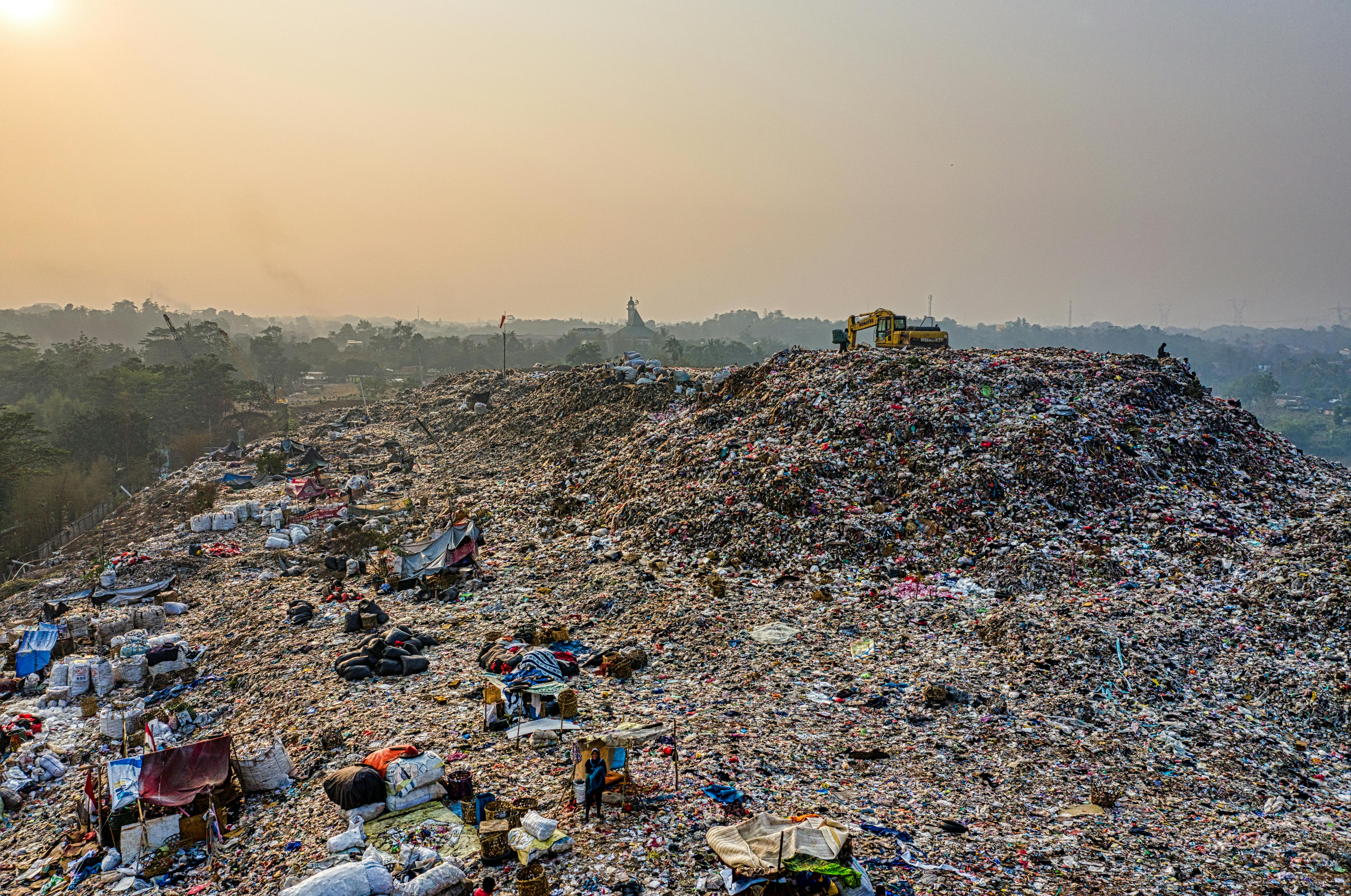 Dirty foggy air in the background, a big hill of piled trash in foreground, on top of it is one excavator 